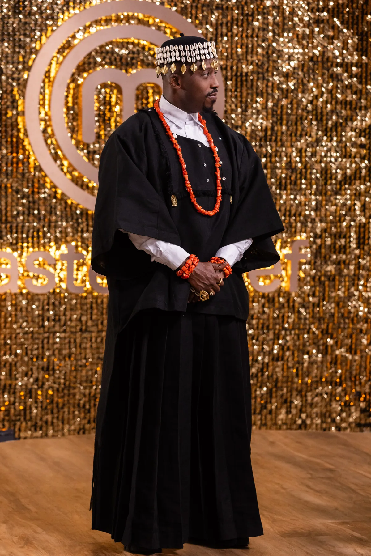 Chef Eros in black Nigerian regalia with coral beads and a cowrie-shell crown, on the MasterChef stage